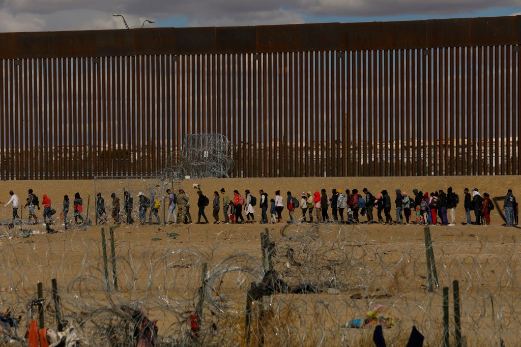 Migrants seeking asylum gather beside the border wall, Texas National Guard stand in the background.